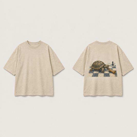 Front and back of a warm-sand oversized tee; a tortoise approaches a fallen chess piece on a small checkerboard patch on the back.